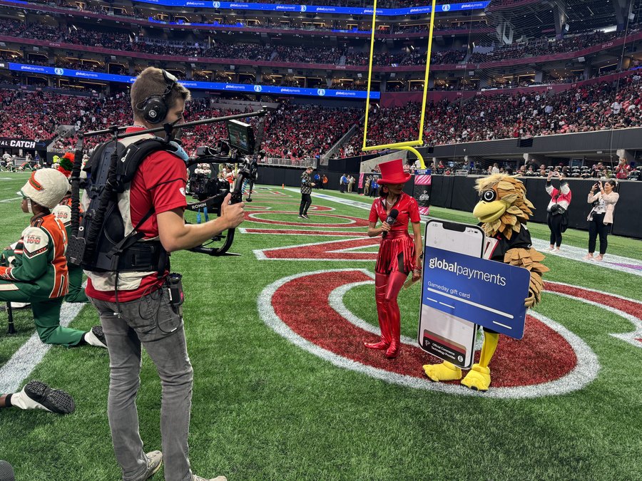 Chelsea running an on-field sponsor segment with the Falcons mascot and a camera operator